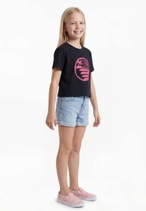 Fille souriante debout portant un t-shirt noir avec un motif plage rose, un short en jean bleu clair et des chaussures slip-on rayées roses sur fond blanc.