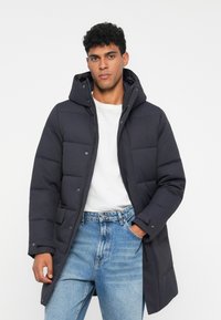 Mørkeblå puffjakke med hætte, der har quiltede paneler, frontknapper og sidelommer, båret over en hvid skjorte og blå jeans.