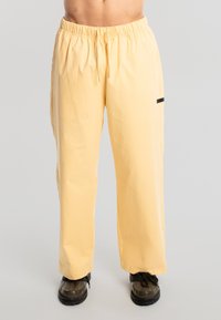 Modelo masculino vistiendo pantalones sueltos de color amarillo pálido con cintura elástica y cordón, combinados con zapatos de senderismo negros, sobre un fondo sencillo.