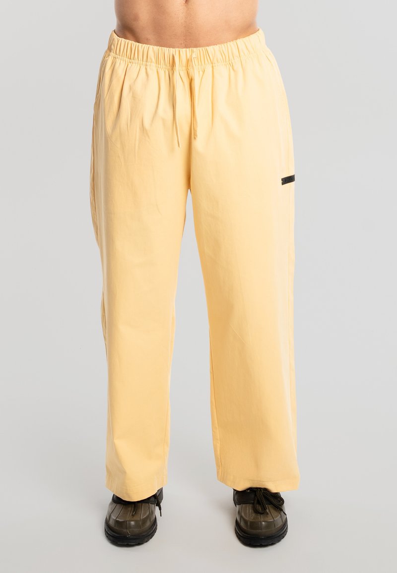 Modelo masculino vistiendo pantalones sueltos de color amarillo pálido con cintura elástica y cordón, combinados con zapatos de senderismo negros, sobre un fondo sencillo.
