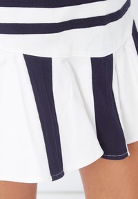 Falda blanca con pliegues verticales en azul marino, llevada por encima de la rodilla, mostrando parte de las piernas.