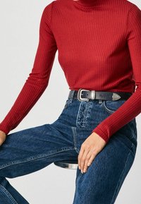 Un haut rouge à manches longues de style côtelé associé à un jean bleu en denim taille haute, souligné par une ceinture en cuir noir avec une boucle en argent.