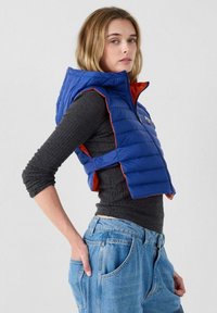 Chaleco acolchado azul con interior naranja, hombros estructurados, cremallera frontal y ajuste holgado, usado sobre una camisa de manga larga gris.