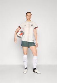 Jersey jalkapalloon Nike, jossa on vaalea beige väri vihreillä ja punaisilla yksityiskohdilla, yhdistettynä vihreisiin shortseihin, valkoisiin sukkiin ja nappulakenkiin.