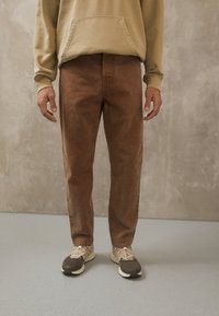Ruske denim-housut med rett snitt, kombinert med en lys beige hettegenser og nøytrale sneakers, mot en teksturert vegg.