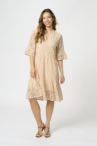 Spetsklänning i ljus persika med trumpetärmar, V-ringning och lager på lager-skirt. Har blommönster och en halvgenomskinlig textur.
