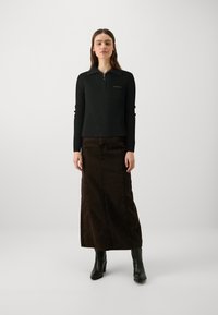 Haut noir côtelé à manches longues avec un zip quart et un détail de logo, associé à une jupe maxi en denim marron et des bottes à talons noires.