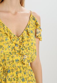 Gul blommig klänning med volangdetaljer på axlarna och halsringningen. Har små blå och rosa blommor på ett strukturerat tyg.