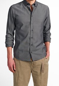 Chemise grise à boutons avec une texture douce, manches longues retroussées jusqu'aux coudes, et petit col, associée à un pantalon beige clair.
