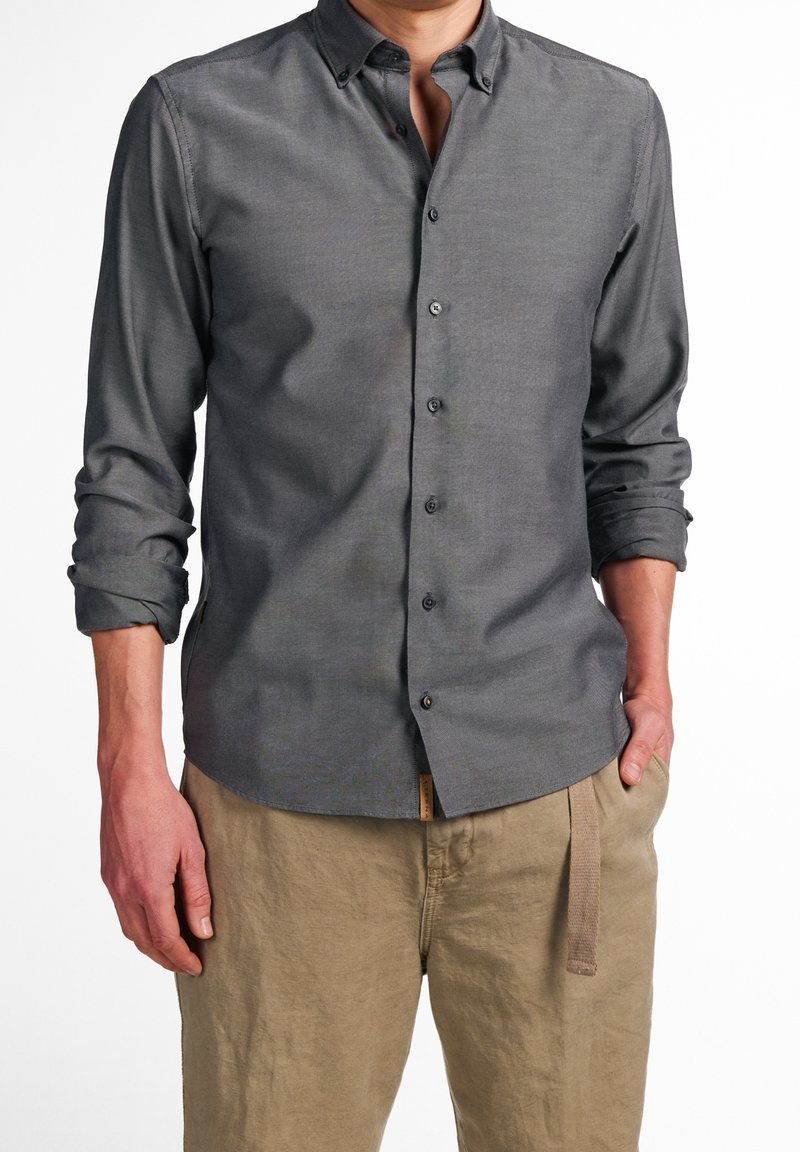 Chemise grise à boutons avec une texture douce, manches longues retroussées jusqu'aux coudes, et petit col, associée à un pantalon beige clair.