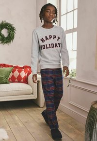 Szary sweter z napisem "HAPPY HOLIDAY" w bordowych literach, w połączeniu z kraciastymi spodniami flannel w odcieniach granatu i czerwieni oraz czarnymi kapciami. Przytulne wnętrze.