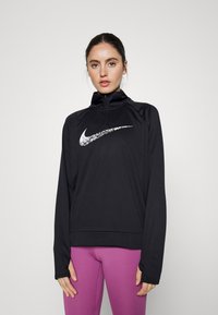 Top sportivo nero a maniche lunghe con zip a quarto, con il logo Nike bianco sul petto. Indossato con leggings viola. Tessuto liscio e elasticizzato.