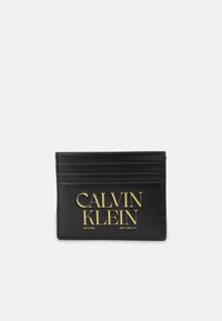 Černý kožený držák na karty má šest slotů, zlatě embosované logo "CALVIN KLEIN" a další text "SINCE 1968 NEW YORK CITY" na dně.