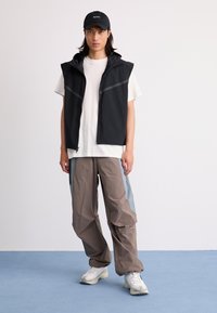 Young man standing, wearing black sleeveless jacket over white T-shirt, two-tone loose pants, white sneakers, and black cap with text.