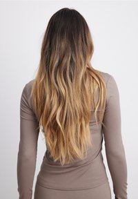 Lunghi capelli ondulati biondo-castano a ombre che scendono lungo la schiena di una persona che indossa un top a maniche lunghe di color taupe aderente e una gonna.