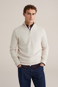 Beige Strickpullover mit Viertelreißverschluss und strukturiertem Muster, mit gerippten Bündchen und Saum, über einem dunklen Hemd getragen und kombiniert mit blauen Jeans.