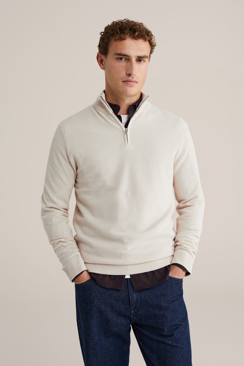 Beige Strickpullover mit Viertelreißverschluss und strukturiertem Muster, mit gerippten Bündchen und Saum, über einem dunklen Hemd getragen und kombiniert mit blauen Jeans.