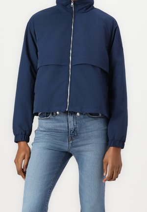 Marineblå lynlåstrøje med høj krave og elastiske manchetter båret sammen med lyseblå, højttaljede jeans i en afslappet stilling.