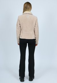 Frau mit langen Haaren, die eine beigen Wildlederjacke mit Schaffellkragen, schwarze Schlaghosen und schwarze Stiefel trägt, vor einem grauen Hintergrund steht.