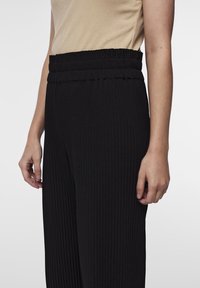 Pantalons noirs côtelés avec une taille élastique, présentant des rayures verticales. Le matériau semble doux avec une texture lisse.