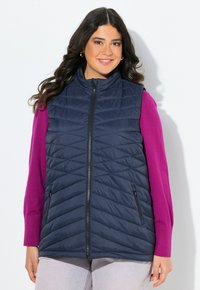 Gilet imbottito navy con pattina, collo alto, zip frontale e tasche laterali, indossato sopra un top a maniche lunghe di un vivace rosa. 