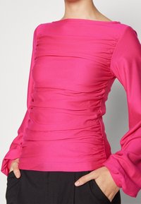 Sittande långärmad topp i klarrosa med rynkad struktur framtill och vida ärmar. Materialet verkar mjukt med en lätt glans.