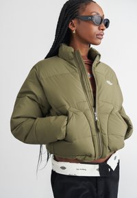 Giacca puffer verde oliva con zip frontale, colletto alto e design trapuntato; indossata sopra un top marrone, abbinata a pantaloni neri e una cintura bianca.