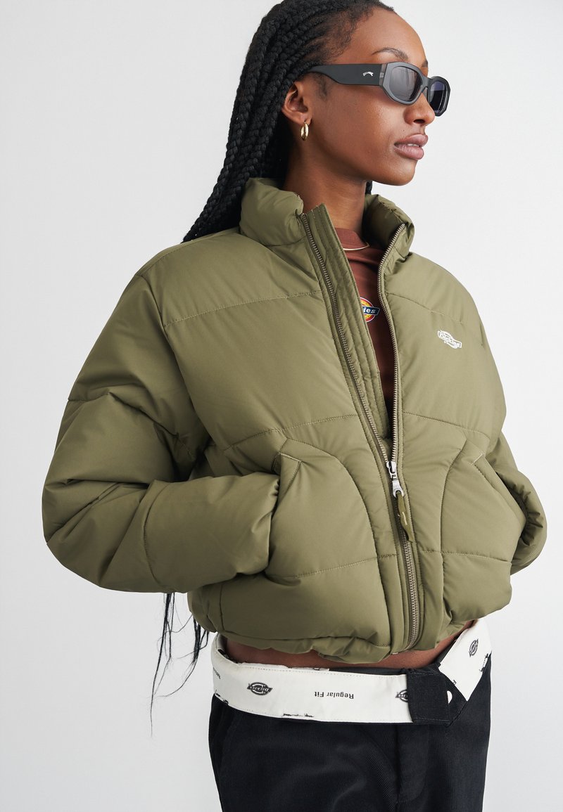 Giacca puffer verde oliva con zip frontale, colletto alto e design trapuntato; indossata sopra un top marrone, abbinata a pantaloni neri e una cintura bianca.
