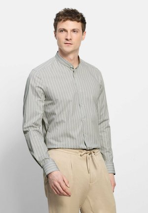 Mann trägt hellgraues, vertikal gestreiftes Hemd mit Stehkragen und beige Hose mit Kordelzug, steht vor schlichter weißer Wand.