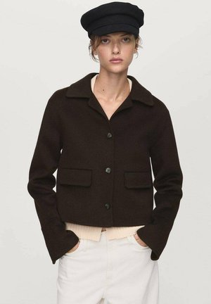 Veste courte marron avec fermeture à boutons, col évasé et deux poches avant. Le modèle porte un haut clair et une casquette noire.