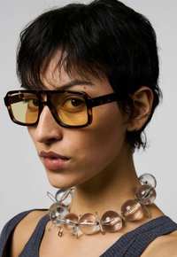 Overmaat tortoise zonnebril met gele lenzen, gecombineerd met een ketting van heldere kralen met metalen accenten, gedragen bij een grijze top met textuur.