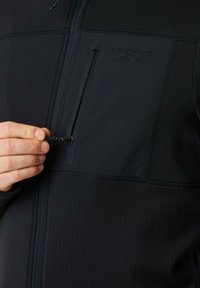 Zwarte jack met rits in een gestructureerde stof, met een borstzak met een waterdichte rits en een reliëflogo. Close-up van een hand die de rits aanpast.