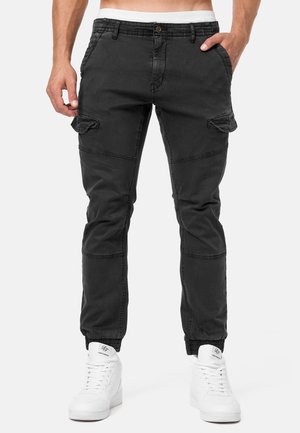 Homme portant un pantalon cargo noir avec des poignets élastiques et des baskets montantes blanches, debout les mains dans les poches.