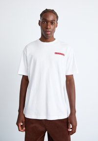 Hvid bomulds T-shirt med korte ærmer, prydet med et rødt rektangulært logo på venstre side. Løs pasform med rund hals.