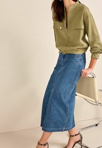 Blusa ligera en verde oliva, con bolsillos en el pecho, cremallera hasta la mitad y un corte relajado, combinada con jeans de mezclilla azul de cintura alta y pierna ancha.