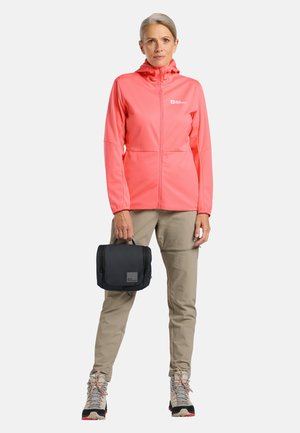 Vrouw draagt een koraalkleurig jack met ritssluiting en kaki broek, houdt een zwarte reistas vast, staand tegen een witte achtergrond.