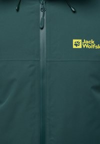 Veste verte avec une texture lisse et mate. Présente une fermeture éclair à l'avant, une capuche avec cordon de serrage ajustable, et un logo jaune sur la poitrine.