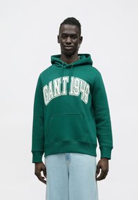 Sudadera verde con capucha, gran texto "GANT 1949" en blanco y un bolsillo frontal. Combinada con vaqueros de pierna ancha en azul claro.