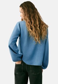 Camisa de mezclilla azul claro de manga larga con un ajuste suelto, cuello redondo y mangas fruncidas, combinada con jeans negros.