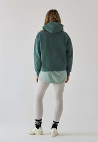 Hoodie w kolorze ciemnozielonym, nałożony na jasnoniebieską koszulę. Białe legginsy, skarpetki w wzorki i kremowe sneakersy dopełniają stylizację.