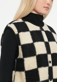 Gilet a scacchi nero e crema realizzato in pile testurizzato con chiusura a bottone, caratterizzato da un design senza maniche e collo rotondo.