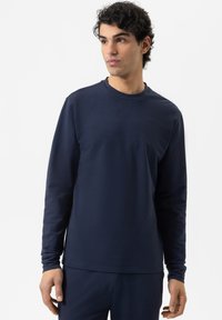 Junger Mann mit lockigem Haar, der ein einfaches, navyblaues Langarmshirt und passende Hose trägt und leicht nach rechts schaut.