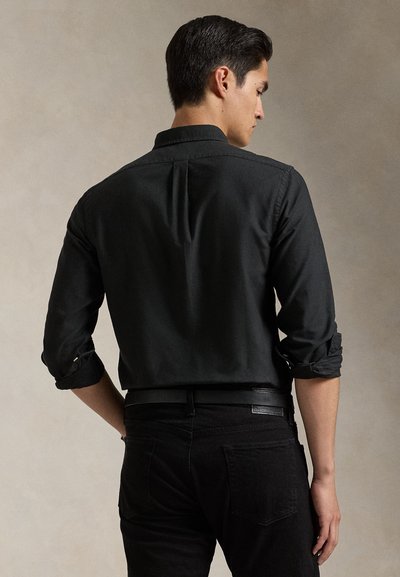 Moški, obrnjen stran, v zavihanem črnem srajci, zataknjen v črne hlače s črnim usnjenim pasom na nevtralnem ozadju.