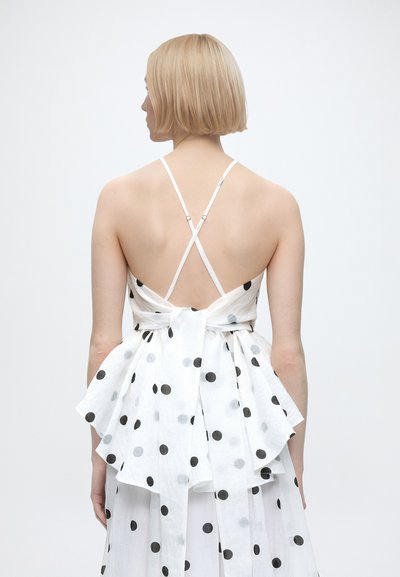 Femme aux cheveux blonds courts portant une robe blanche à pois noirs, bretelles fines croisées et un grand nœud dans le bas du dos.