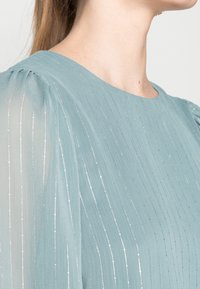 Błękitna bluzka z pionowymi srebrnymi paskami, z okrągłym dekoltem i miękkimi, marszczonymi rękawami. Lekki, przezroczysty materiał o delikatnym połysku.