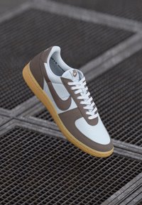 Bijela i smeđa Nike tenisica s teksturiranim gornjim dijelom, ravnim vezicama i gumiranim potplatom, koja prikazuje kontrastni dizajn i detalj s logotipom.