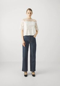 Kvinne som har på seg en hvit blondebluse med kort erm, mørkeblå høyt midjesittende vide jeans og svarte Mary Jane-sko, som står mot en ensfarget bakgrunn.