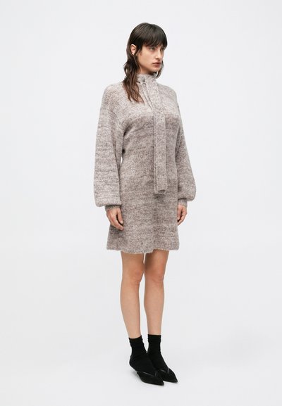 Pletené šaty v jemnej béžovej farbe s imitáciou goliera, dlhými rukávmi a voľným strihom; majú jemnú textúru a rebrované detaily na manžetách.