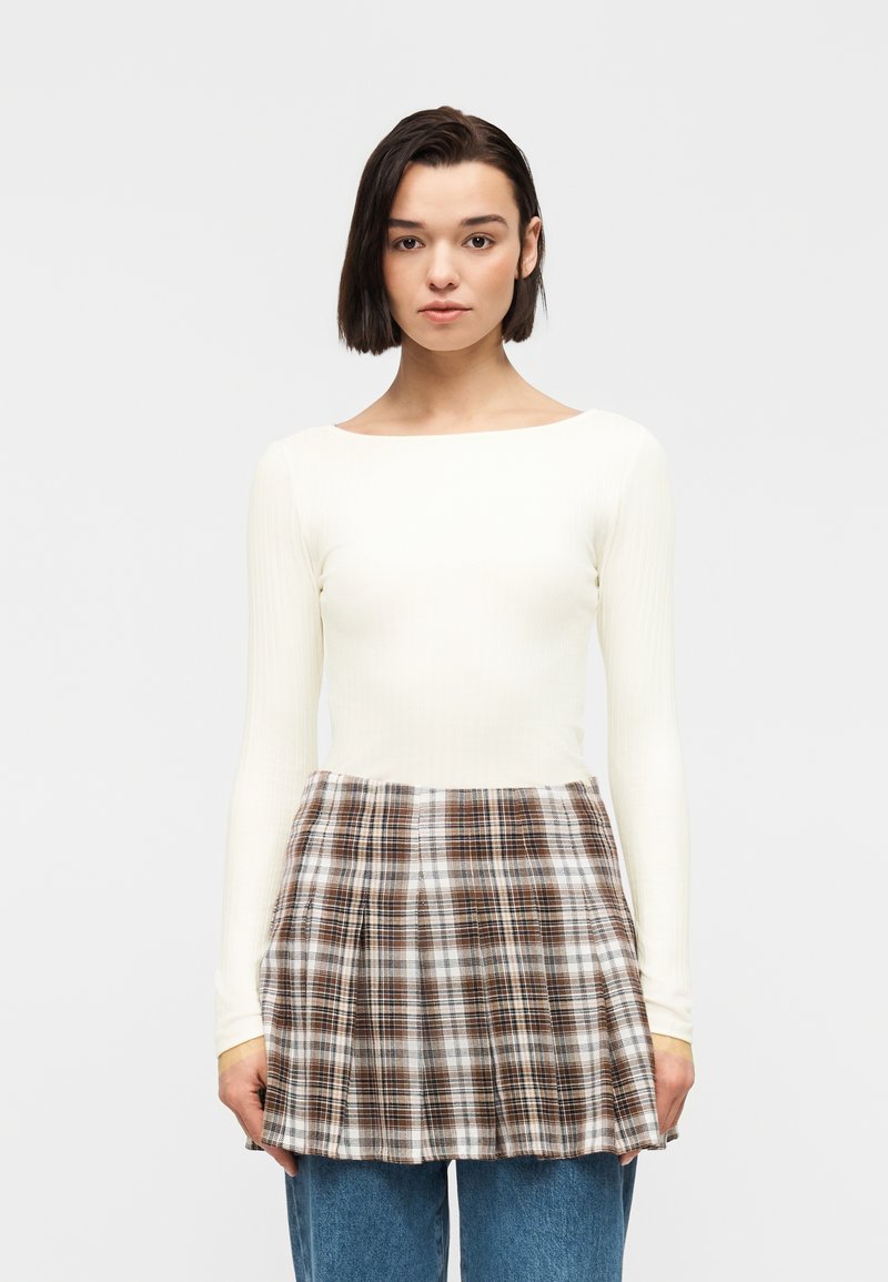 Νέα γυναίκα με κοντά σκούρα μαλλιά, ντυμένη με λευκό μπλουζάκι μακρυμάνικο και καρό καφέ ποδιά πάνω από μπλε τζιν, στέκεται μπροστά σε απλό φόντο.