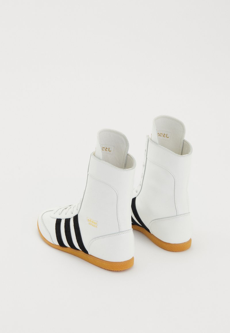 Weiße High-Top-Sneaker mit schwarzen Streifen, Gummisohle, strukturiertem Leder-Obermaterial und goldener Markenprägung an der Seite. Entwickelt für sportliche Zwecke.